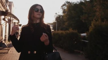 Şık siyah elbiseli ve güneş gözlüklü güzel kız gün batımında bir şehir caddesinde duruyor. Moda konsepti. Yavaş çekim