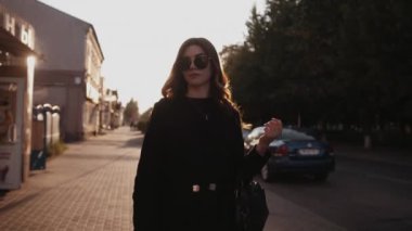 Güneş gözlüklü modaya uyan genç bir kız gün batımına karşı şehir caddesinde yürüyor. Moda ve tarz kavramı. Yavaş çekim