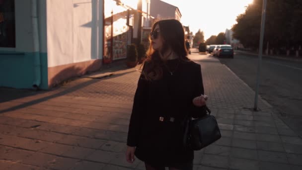 Ein hinreißendes junges Mädchen in schwarzer Kleidung und Sonnenbrille steht bei Sonnenuntergang auf einer Straße in der Stadt und glättet ihre Haare. Zeitlupe — Stockvideo