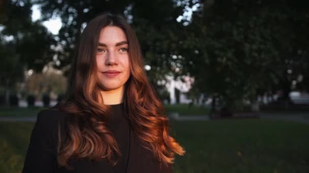 Portrait of a charming young girl with gorgeous long hair standing against the backdrop of trees at sunset. Slow motion — Stock Video