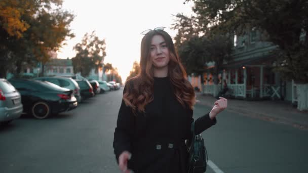 Mladá dívka ve stylových černých šatech jde po silnici podzimního města a obrací se k fotoaparátu s třepetajícími se vlasy. Zpomalený pohyb — Stock video