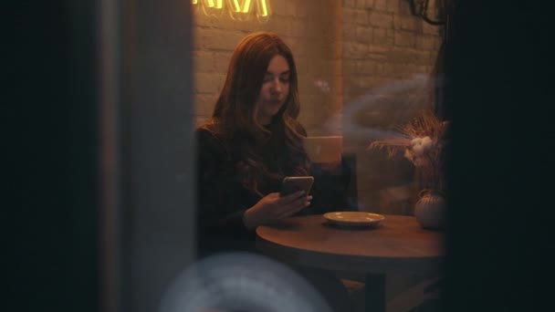 Красива молода дівчина п'є каву в кафе і використовує мобільний телефон. Вид через вікно. Повільний рух — стокове відео