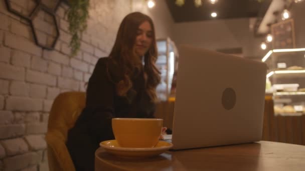 Красива молода дівчина з солодкою посмішкою сидить в кафе з оригінальним інтер'єром і працює в ноутбуці. Повільний рух — стокове відео