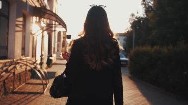 Şık siyah elbiseli enerjik bir genç kız gün batımının arkaplanına doğru gidiyor ve kameraya gülümsüyor.