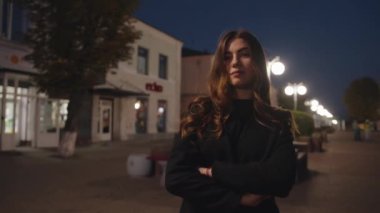 Göz kamaştırıcı genç bir kız, gecenin geç saatlerinde bir yaya caddesinde kollarını göğsüne kavuşturarak ayakta duruyor. Yavaş çekim