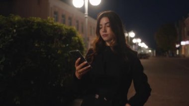 Güzel bir genç kız şehrin akşam caddesinde duruyor ve bir cep telefonunun ekranında gülüyor.