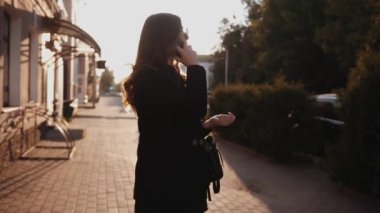 Şık siyah elbiseli ve güneş gözlüklü güzel bir kız gün batımında şehir sokaklarında telefonla konuşuyor. Moda konsepti. Yavaş çekim