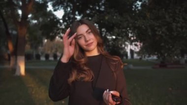 Siyah şık elbiseli güzel bir kızın portresi bir şehir caddesinde ağaçların arasında duruyor ve gülüyor. Yavaş çekim