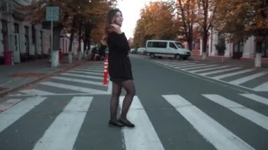 Şık siyah ceketli ve kısa etekli genç bir kız sarı sonbahar ağaçlarıyla karşıdan karşıya geçiyor. Yavaş çekim