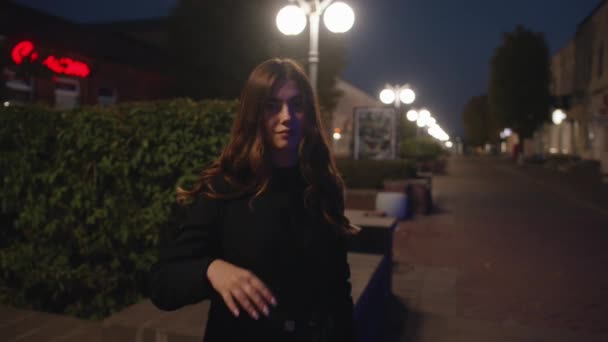 Una hermosa joven se levanta tarde por la noche en una zona peatonal de la ciudad con letreros de neón en los edificios y endereza su lujoso cabello. Movimiento lento — Vídeo de stock