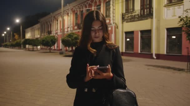 Trendy jonge meid staat tegen de achtergrond van de historische gebouwen van de avondstad en bladert door het scherm van een telefoon. Langzame beweging — Stockvideo