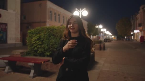 Roztomilá mladá dívka ve stylovém černém oblečení jde podél večerní ulice a má výhled na město. Zpomalený pohyb — Stock video