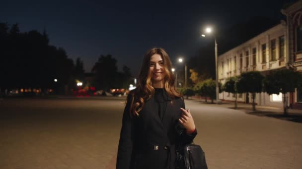 A charming young girl in a stylish black outfit is standing on a night street lit by lanterns and smiling. Slow motion — Stock Video