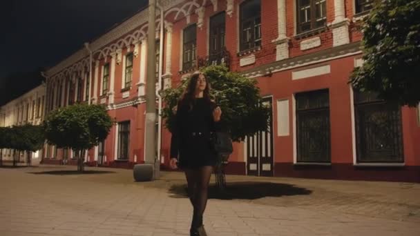 Długowłosy młoda dziewczyna w czarnym krótkim stroju idzie wieczorem na tle zabytkowego budynku. Kąt fotografowania dolnego — Wideo stockowe