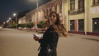 Muhteşem genç bir kız, akşam şehrin tarihi köşesinden içeri giriyor ve telefon ekranından yapraklar dökülüyor. Yan görünüm