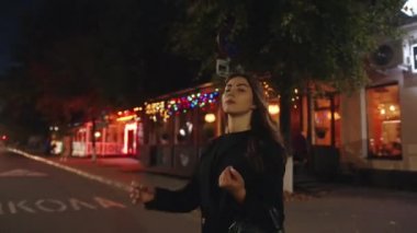 Şehvetli genç bir kız, renkli çelenklerin ve neon ışıklarının olduğu bir yaya geçidinin yanında duruyor. Yavaş çekim