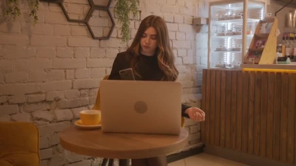 Egy komoly fiatal lány ül egy kávézóban, szemben a háttérben egy fehér téglafal díszített természetes növények és dolgozik egy laptop. Lassú mozgás. — Stock videók