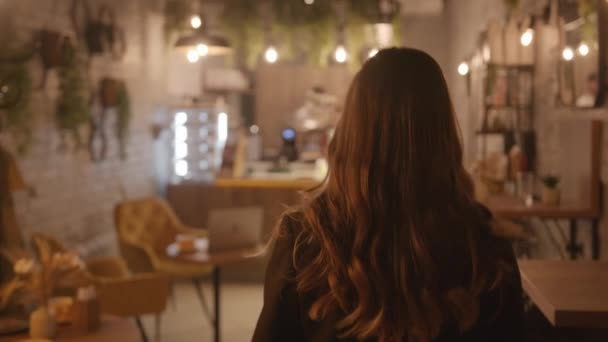 Красивая девушка проходит по коридору кафе, украшенному живыми растениями, и садится за стол с ноутбуком — стоковое видео