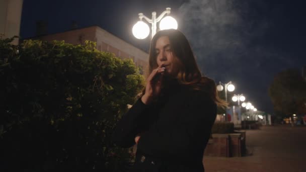 Elegante chica joven libera una nube de un vapor en la calle de la noche y se para en una bocanada de humo. Movimiento lento — Vídeos de Stock