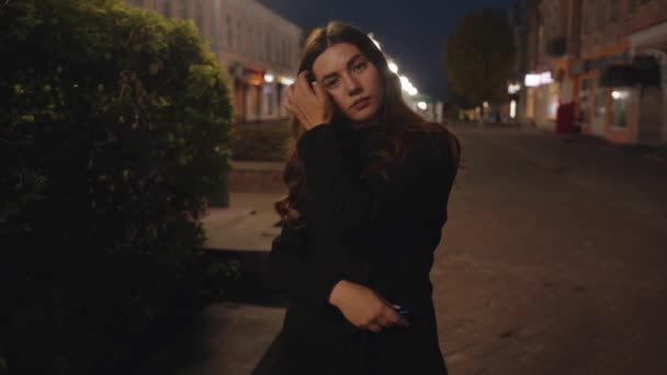 Uma jovem glamourosa fica de pé com os braços cruzados em seu peito em uma rua pedonal da cidade no final da noite. Movimento lento — Vídeo de Stock