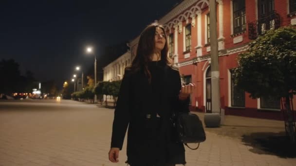 Een elegant jong meisje wandelt langs de verlichte voetgangerszone van de avondstad en geniet van een prachtig uitzicht — Stockvideo
