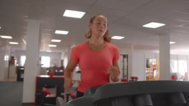 Сфокусированная молодая девушка в розовой футболке тренируется в спортзале и бежит на беговой дорожке. Понятие здорового образа жизни. Медленное движение — стоковое видео