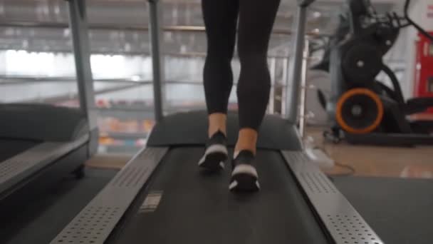 Närbild av kvinnliga ben i svarta sneakers och svarta leggings på ett löpband i gymmet. Bakåt. Långsamma rörelser — Stockvideo