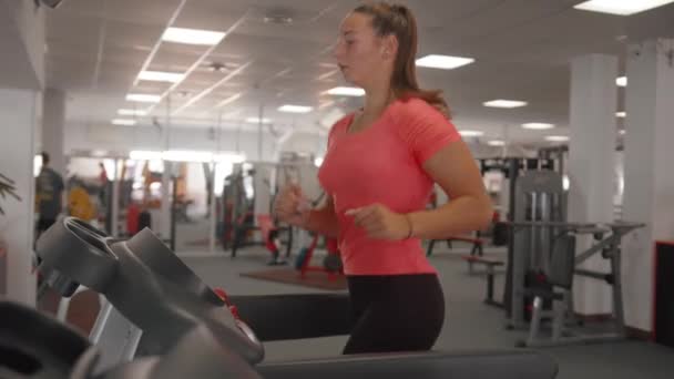 Красивая молодая девушка в розовой футболке тренируется в тренажерном зале на беговой дорожке, чтобы сжечь калории и укрепить мышцы — стоковое видео