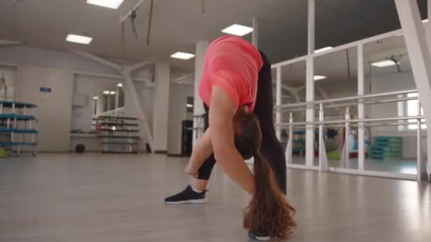 Een mooi jong meisje strekt zich uit na krachttraining in de sportschool en maakt voorwaartse bochten met een twist van het lichaam — Stockvideo