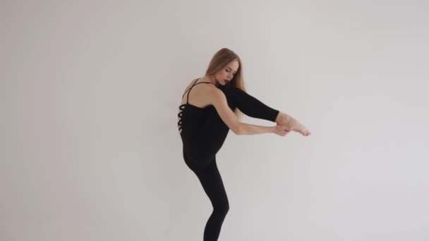 Une jeune fille avec un maquillage lumineux dans un beau haut noir et leggings fait étirement dans un studio blanc et soulève sa jambe haute — Video