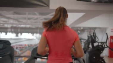 Terli tişörtlü genç bir kadın spor salonunda kardiyo eğitimi alıyor ve vücut dayanıklılığını arttırmak için koşu bandında koşuyor. Arka plan