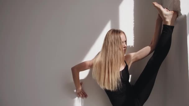 Leende ung flicka gör stretching och sätta sitt ben på väggen och står i en vertikal split lutande på väggen — Stockvideo