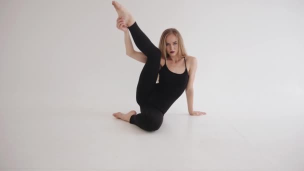 Uma jovem espetacular em um top preto e leggings está envolvida em alongamento no estúdio e se senta em um cordel com a perna dobrada à cabeça — Vídeo de Stock