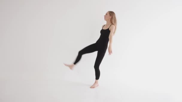 Een mooi jong meisje in een zwarte top en leggings is bezig met stretching in de studio en doet een verticale split — Stockvideo