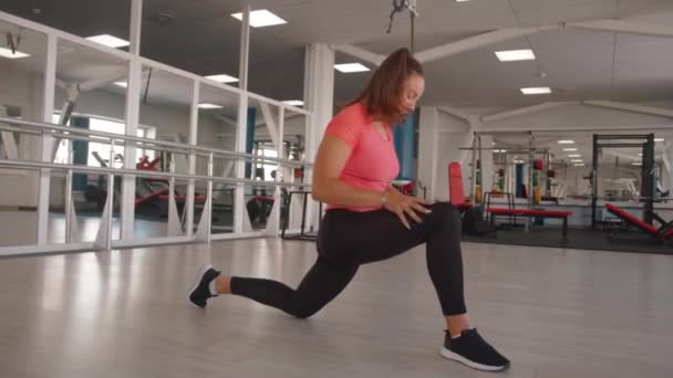 Молодая женщина в розовой футболке тренируется в современном спортзале и делает статические глубокие выпады. Концепция силы и мотивации — стоковое видео