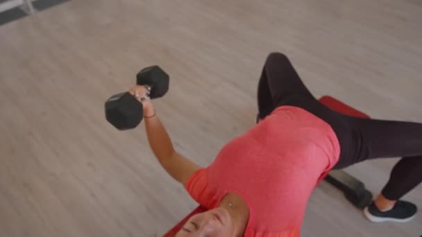 Uma jovem com uma camiseta rosa e perneiras pretas treinando no ginásio e faz supino com halteres. Vista de cima — Vídeo de Stock