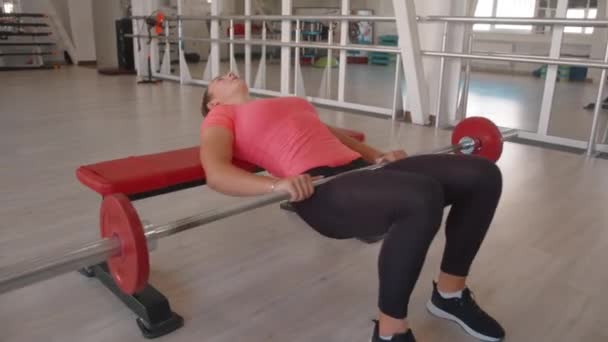 Молодая девушка в спортзале делает упражнение, чтобы поднять таз на скамейке с штангой и накачивает ягодицы и мышцы задней части бедра — стоковое видео
