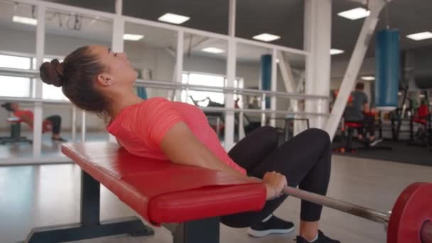 Junge Frau im Fitnessstudio führt ein Beckenheben auf einer Bank mit einer Langhantel durch und schüttelt die Muskeln des hinteren Oberschenkels und des Gesäßes — Stockvideo