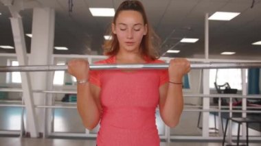 Pembe tişörtlü enerjik genç bir kız spor salonundaki aynanın önünde dikilirken halter kaldırıyor.