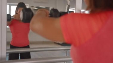 Genç bir kız spor salonunda, önünde duran dambıllarla el kaldırıyor. Aynanın önünde, kaslarını çalıştırmak için.