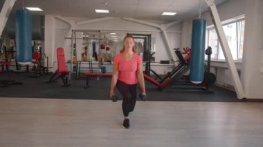 Pembe tişörtlü, siyah tozlu tatlı bir kız spor salonunda antrenman yapıyor ve elinde dambıllarla atlıyor. Yavaş çekim