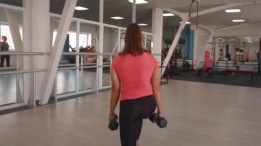 Genç bir kız, spor salonunda hareket halindeki halterlerle hareket ederek bacak ve kalça kaslarını sallıyor. Arka plan