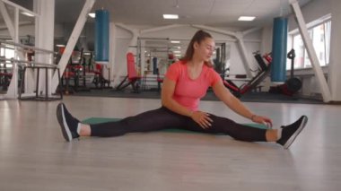 Genç bir kız spor salonunda çalışıyor ve bir paspasın üzerinde otururken esneme hareketleri yapıyor ve geniş bacaklara eğiliyor.