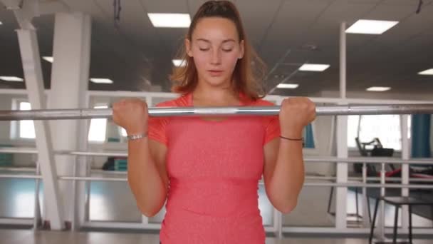 Chica joven y enérgica en una camiseta rosa haciendo levantamiento de pesas para bíceps mientras está de pie sobre el fondo de un espejo en el gimnasio — Vídeos de Stock