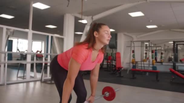 Chica joven atlética en una camiseta rosa y leggins negros de entrenamiento en un gimnasio de moda y hace una fila de pesas con una inclinación — Vídeo de stock