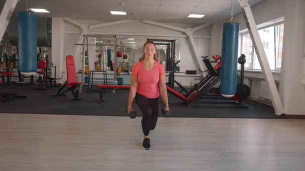 Ένα χαριτωμένο νεαρό κορίτσι σε ένα ροζ T-shirt και μαύρο κολάν κατάρτισης στο γυμναστήριο και κάνει lunges με αλτήρες στα χέρια της. Αργή κίνηση — Αρχείο Βίντεο