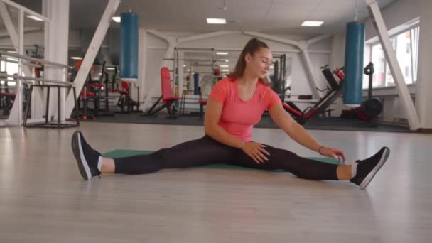 한 소녀 가 체육관에서 운동을 하고 매트 위에 앉아 다리를 넓게 펴고 스트레칭을 합니다 — 비디오