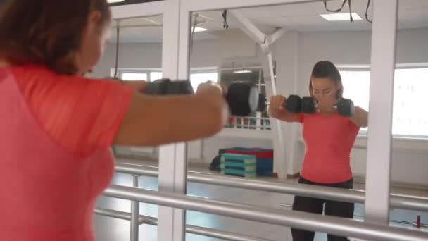 Güzel bir genç kız spor salonunda aynanın önünde antrenman yapıyor ve önünde dambıllarıyla el kaldırıyor. — Stok video