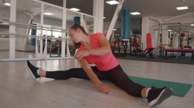 Hoş bir genç kız spor salonundaki bir paspasın üzerinde bacaklarını ayırarak oturur ve gövdesini bükerek bacağına doğru eğilir. Yavaş çekim