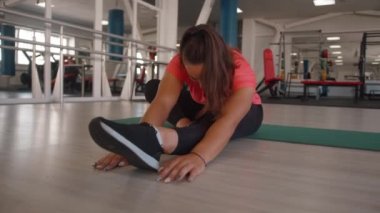 Güzel bir genç kız spor salonunda antrenman yapıyor ve paspasın üzerinde otururken bacağına eğiliyor. Yavaş çekim